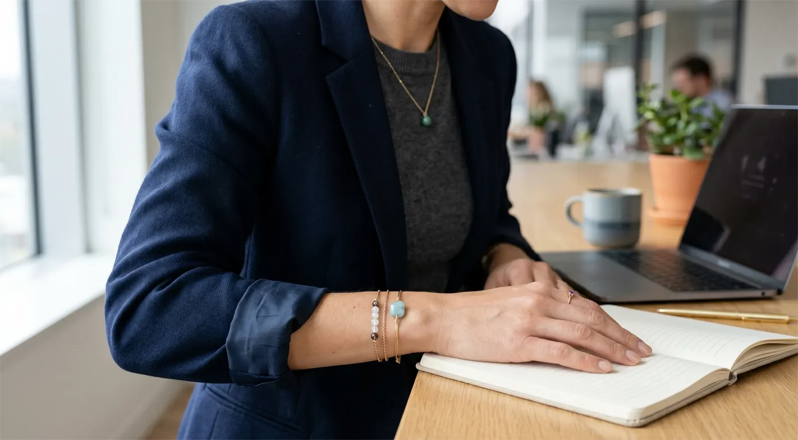Chi tiết trang phục công sở với tay áo blazer, vòng tay đá trung tính, dây chuyền mảnh và nhẫn đơn giản tại bàn làm việc