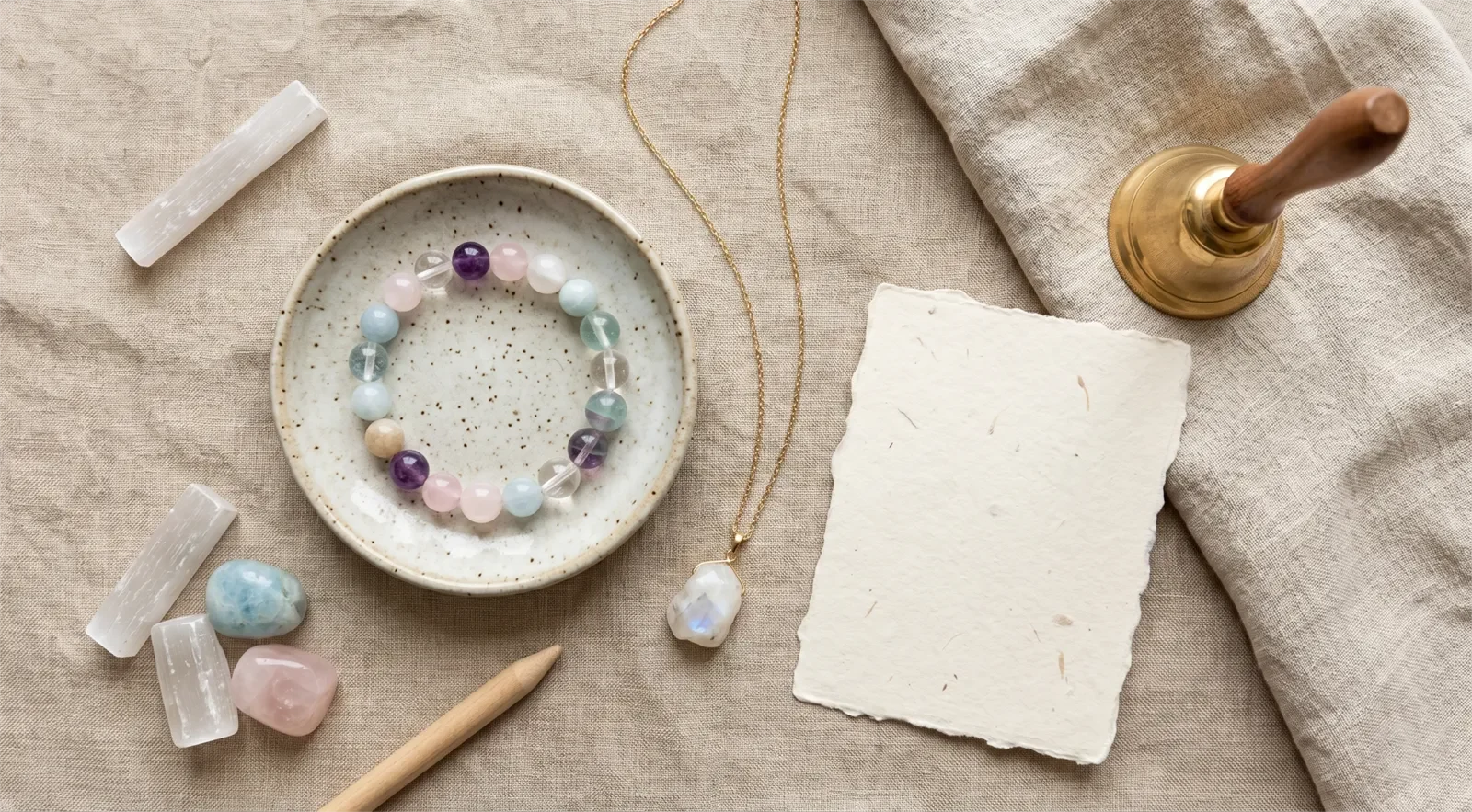 Crystal bracelet and pendant necklace arranged with a bell, linen cloth, ceramic dish, and blank intention card for symbolic cleansing